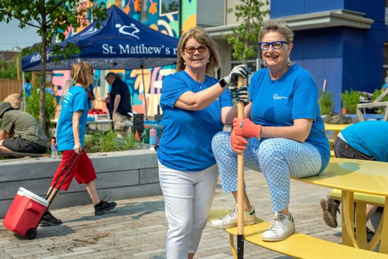 Two elderly women outdoors in blue t-shirts smiling as they hold a rake