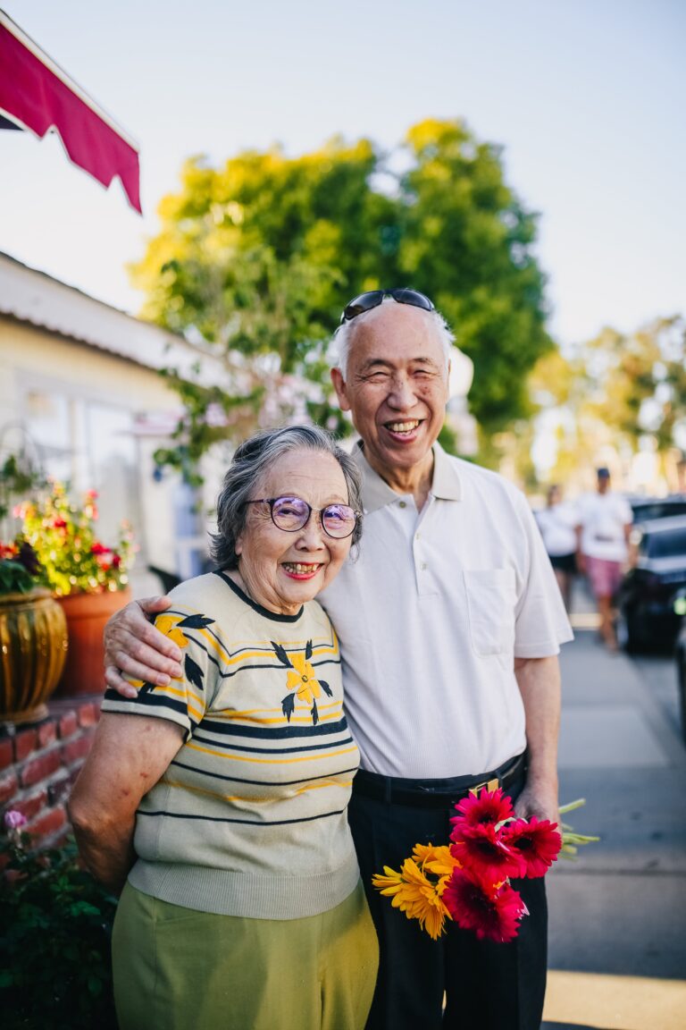 A happy elderly man places right his arm around his spouse's shoulder while holding flowers in his left hand, outside on a sunny day.